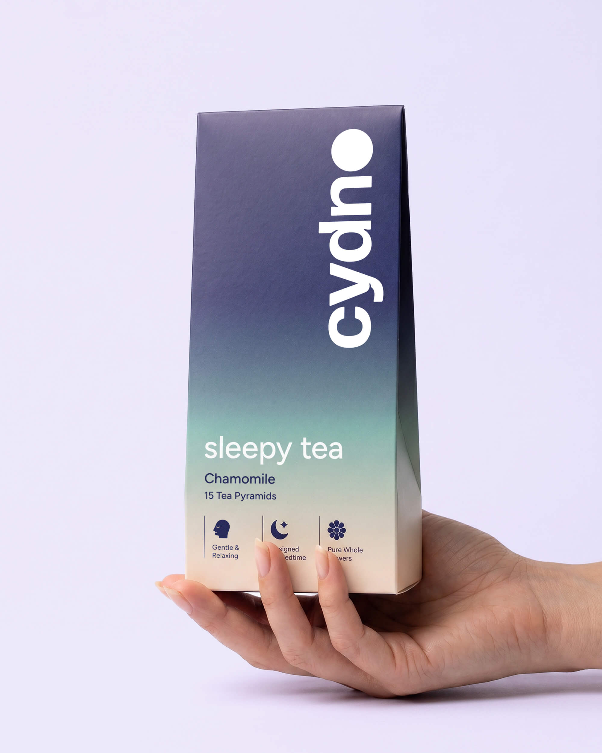 Hand holding an upright Cydno Chamomile carton with its navy-to-teal gradient against lilac