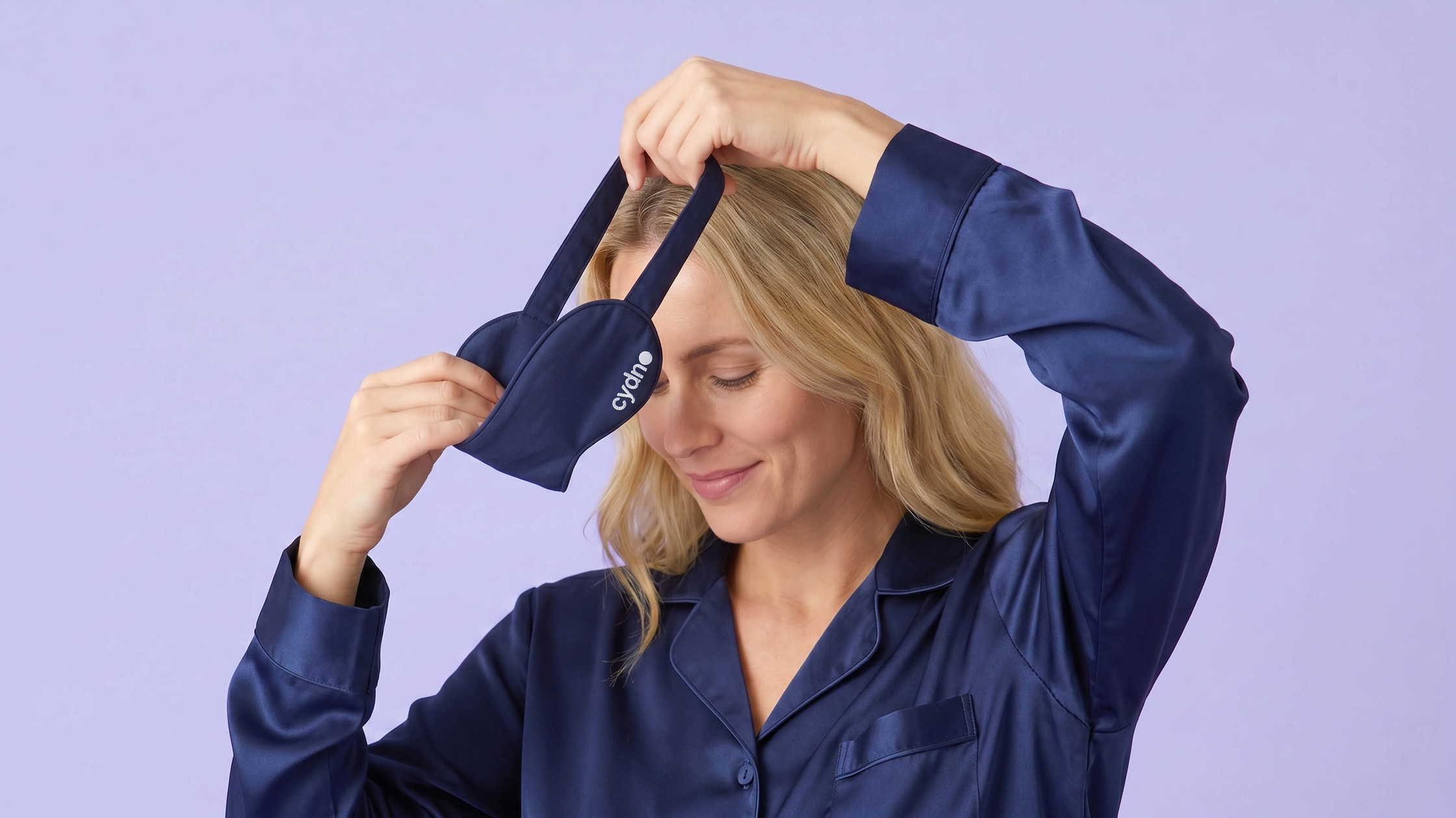 Woman in navy satin pyjamas holding a navy Cydno sleep eye mask with white logo